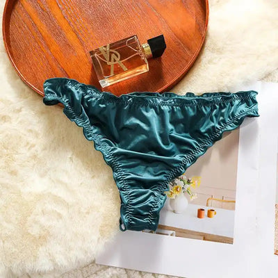 Single dark teal satin thong with ruffled edges laid flat next to a wooden tray and perfume bottle