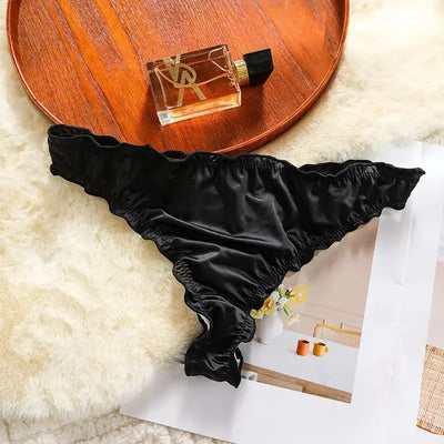 Single black satin thong with ruffled edges laid flat on fur next to perfume bottle