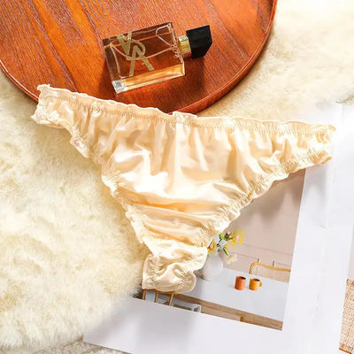 Single ivory satin thong with ruffled edges on fur next to a perfume bottle on wooden tray