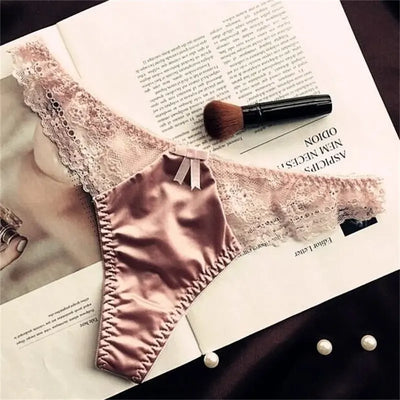 Blush pink satin thong panties with scalloped lace trim and bow, flat lay on magazine with pearls