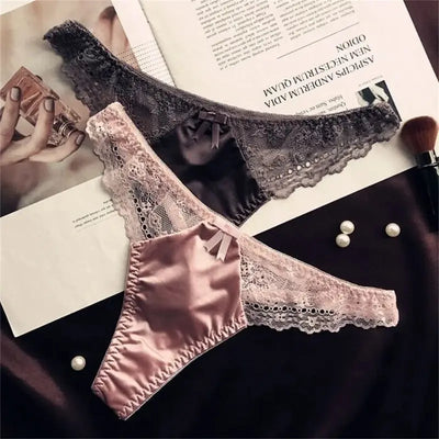 Pink and gray satin thong panties with lace trim and small bow, flat lay on dark fabric with pearls and magazine