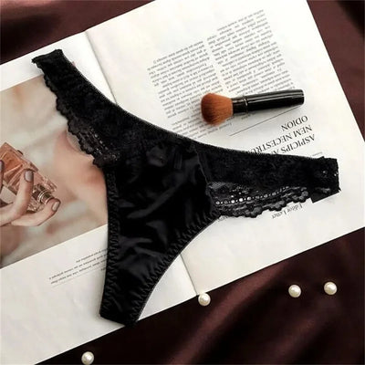 Black satin thong panties with scalloped lace waistband and rhinestone trim, flat lay on open magazine
