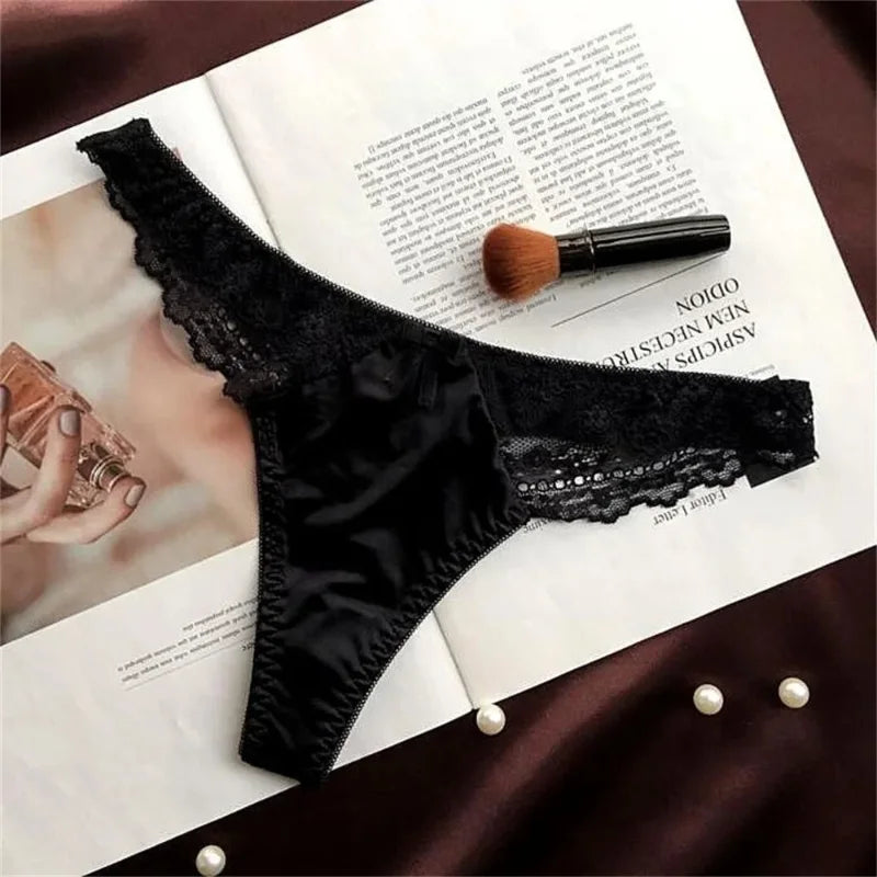 Pink and gray satin thong panties with lace trim and small bow, flat lay on dark fabric with pearls and magazine