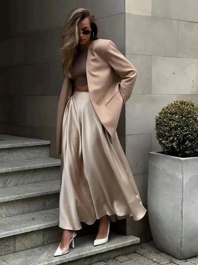 Woman in champagne satin midi skirt and blush blazer posed on stone steps looking down
