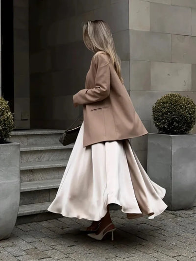 Back view of woman in champagne satin midi skirt and camel blazer walking on cobblestone