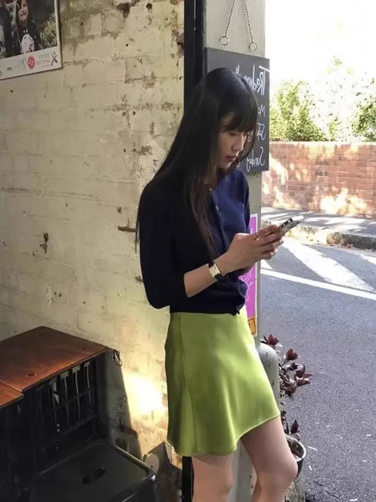 Woman in olive green satin mini skirt and navy top checking phone outdoors