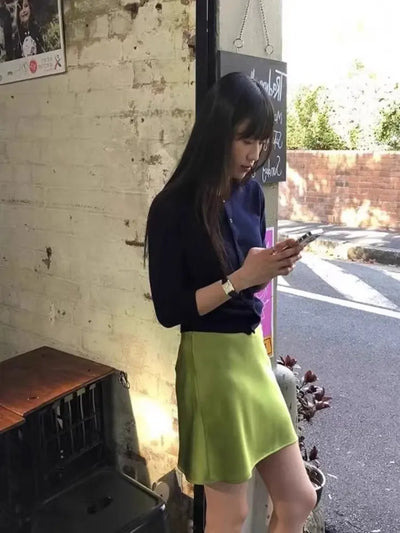 Woman in olive green satin mini skirt and navy top checking phone outdoors