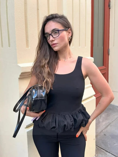 Woman in black bubble-hem peplum top with black pants holding small patent leather bag outdoors