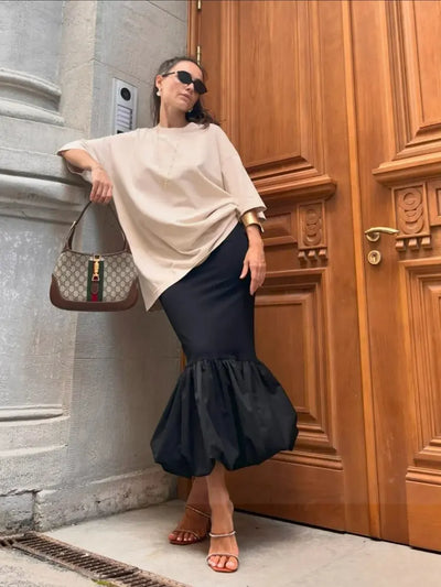 Woman leaning against wooden door in black bubble-hem midi skirt with beige oversized top and Gucci bag