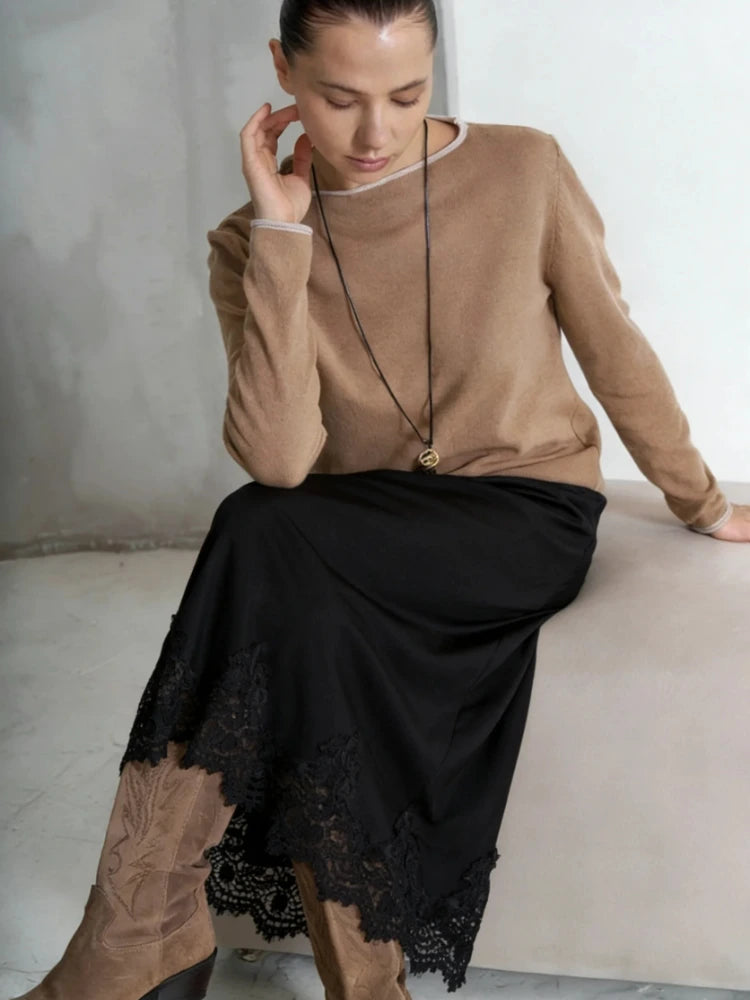 Woman seated wearing a black midi satin skirt with scalloped lace hem, camel knit sweater, and tan cowboy boots
