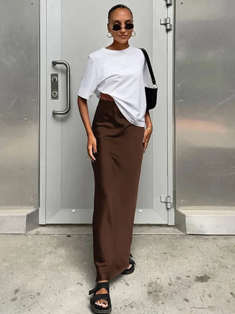 Woman wearing a chocolate brown high-waist satin maxi skirt with a white oversized tee and black sandals