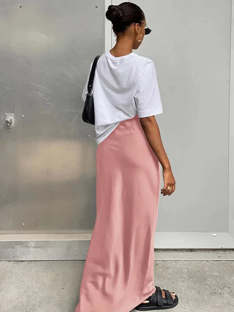 Woman wearing a pink floor-length satin maxi skirt with a white oversized crew-neck tee, black sandals, and a black shoulder bag, viewed from behind against a gray metal wall.