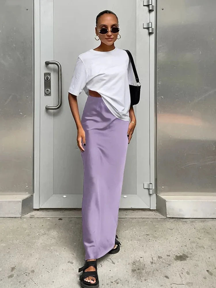 Woman wearing a lavender high-waist satin maxi skirt with a white oversized tee, black sandals, and a crossbody bag, standing in front of a metal door.