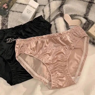 Flat lay of black and blush pink ruched satin panties with Dimple script embroidery on white bedding