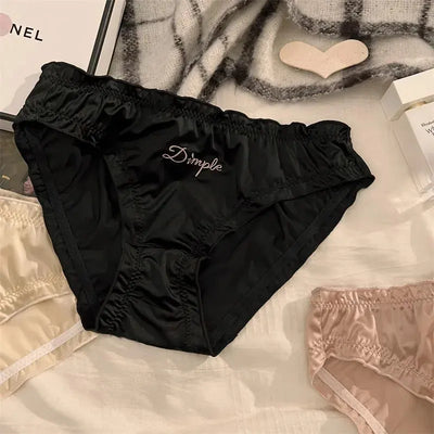 Black ruched satin Dimple panties laid flat next to blush and cream styles on white linen