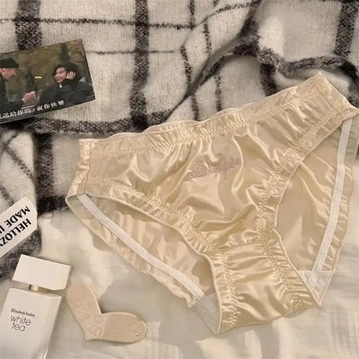 Cream ruched satin Dimple panties with script embroidery displayed on white bedding with accessories