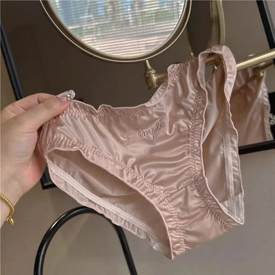 Hands holding blush pink ruched satin Dimple panties showing leg opening and whipstitch finish in mirror