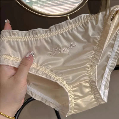 Hands holding cream ruched satin Dimple panties showing front panel and embroidered script detail
