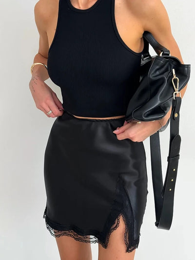 Front view of a black satin mini skirt with lace trim hem and front split worn with a black crop tank top and leather bag