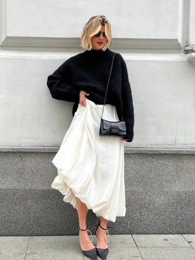 Woman in ivory satin maxi skirt and black turtleneck sweater holding skirt fabric sideways outdoors