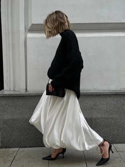 Side profile of woman walking in ivory satin maxi skirt with black pointed-toe heels and chain bag