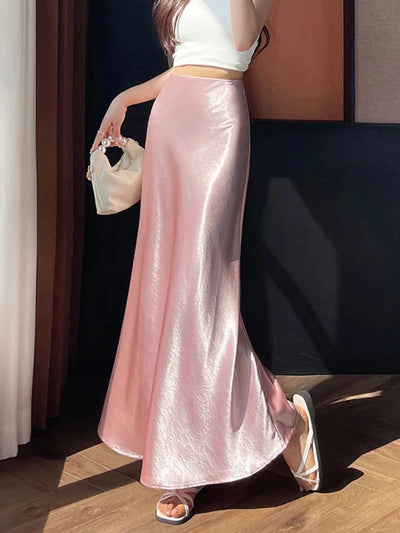 Woman wearing a glossy pink high-waist flared maxi skirt with a white crop top and white sandals, holding a small pearl-handle bag, side view in natural sunlight.