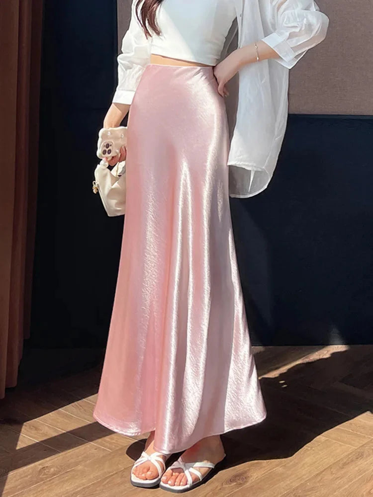Woman wearing a glossy pink high-waist flared maxi skirt with a white crop top and white sandals, holding a small pearl-handle bag, side view in natural sunlight.