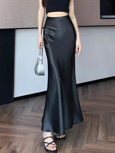 Front view of a black crinkled high-shine fishtail maxi skirt paired with a silver handbag and black strappy sandals