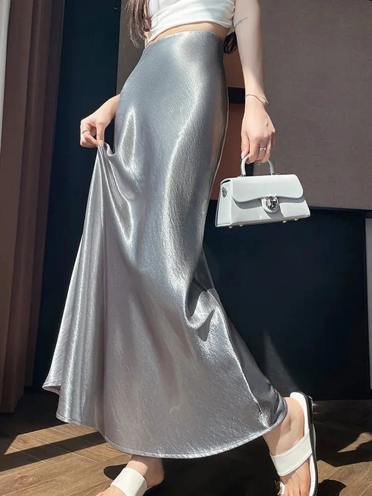 Close-up of a silver crinkled high-shine fishtail maxi skirt held at the hem by a woman in white sandals