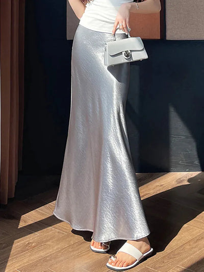 Side profile of a woman in a silver fishtail maxi skirt and white tank top holding a small structured bag