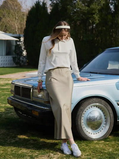 Model in sage green maxi skirt with white long-sleeve top and sneakers leaning against blue car