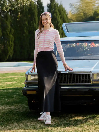 Woman in pink striped cardigan and black satin maxi skirt leaning against a dark vintage car outdoors