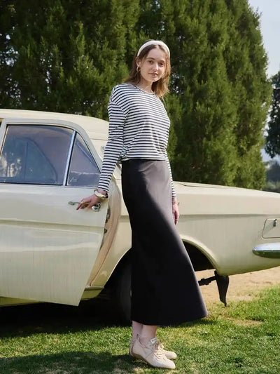 Woman in navy striped top and black satin maxi skirt shown from the side next to a cream vintage car