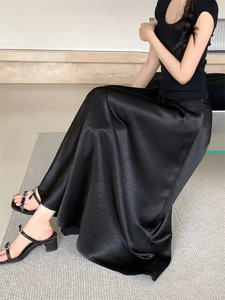 Woman seated wearing a black A-line crinkled satin midi skirt with strappy sandals