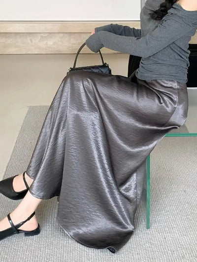 Woman seated holding black quilted bag wearing crinkled gray satin midi skirt with black Mary Jane flats