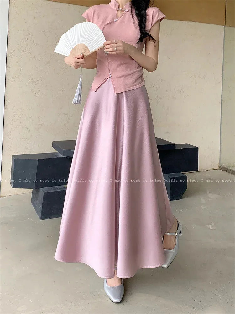 Woman holding a white folding fan wearing a pink flared maxi skirt and matching qipao-collar top with silver Mary Jane heels