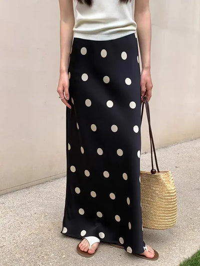Front view of a black polka dot maxi skirt styled with a white knit top and a round straw bag