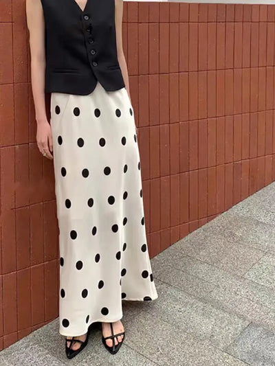 Ivory polka dot maxi satin skirt with glossy finish worn with a black vest against a brick wall