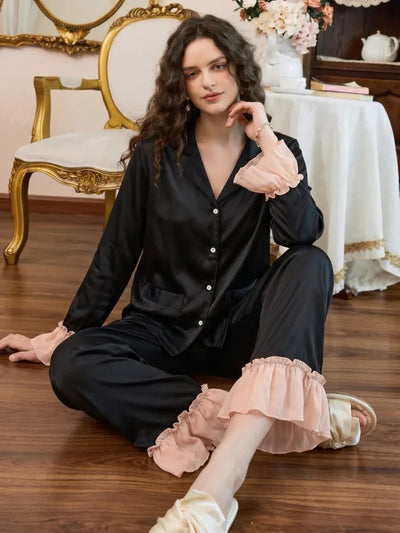 Woman seated in black satin PJ set with pink ruffle cuffs and ankle trim on wood floor