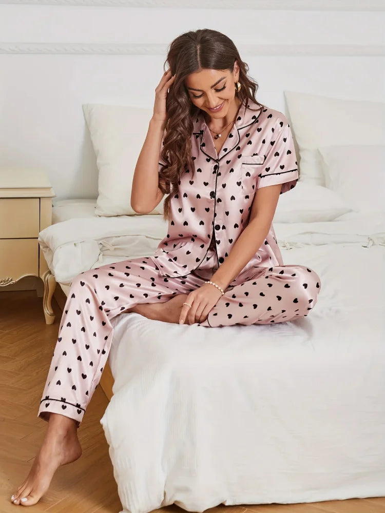 Woman sitting on bed wearing pink satin heart print pajama set with long pants, front view
