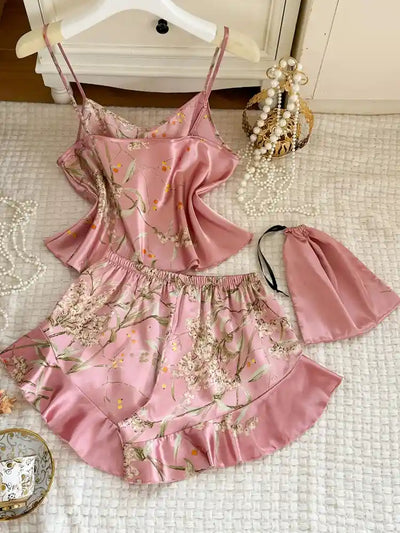 Pink floral satin pajama set with ruffled shorts, cami top, and drawstring pouch on a textured surface