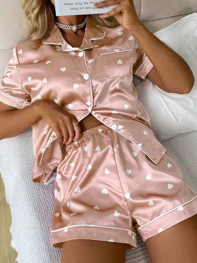 Model wearing blush pink heart-print satin PJ set, seated on white bedding holding a book