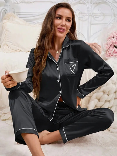 Woman sitting on bed holding a cup, wearing a black satin long-sleeve pajama set with white heart embroidery on the chest pocket