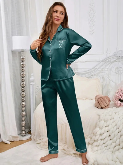 Woman standing in teal satin long-sleeve pajama set with heart embroidery, full front view in bedroom setting
