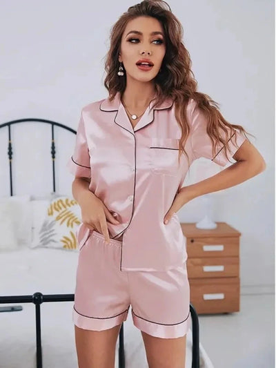 Woman wearing blush satin short-sleeve button-down pajama set with black contrast trim, standing in a bedroom