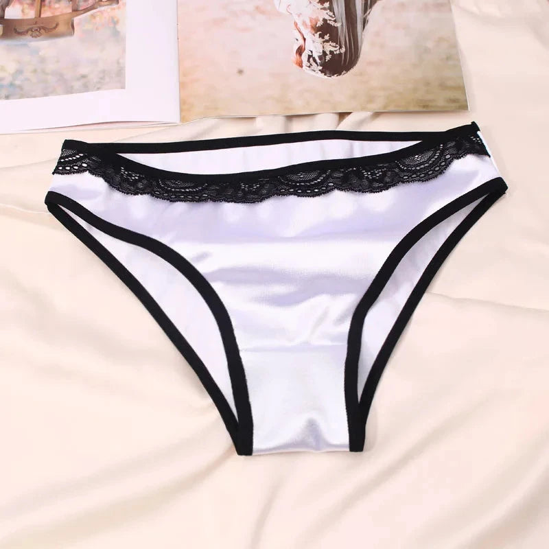 White satin bikini panties with black scalloped lace waistband and contrast leg binding, flat lay on cream fabric