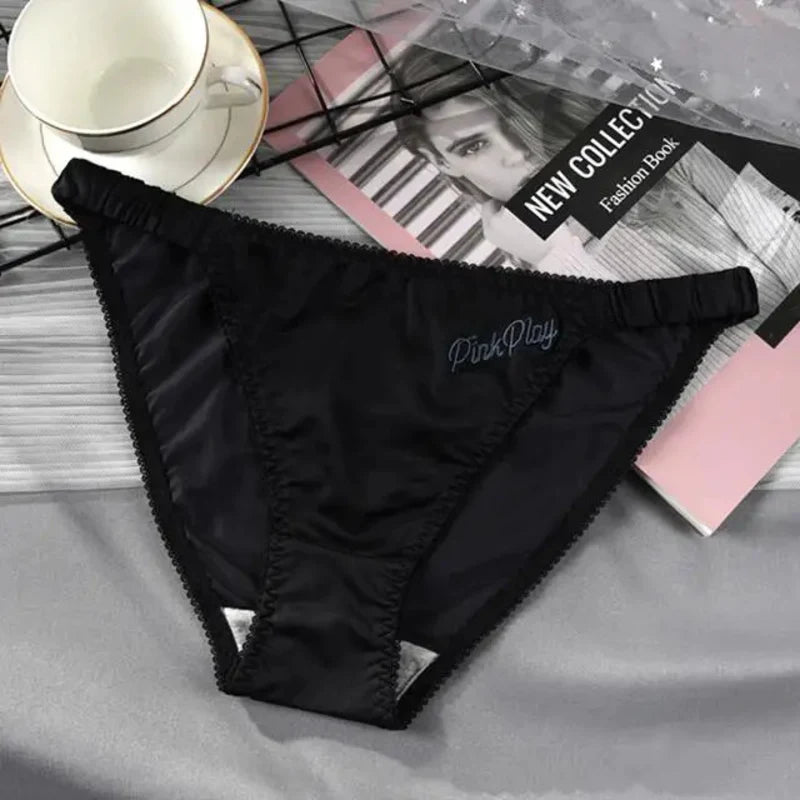 Black satin bikini panties with scalloped trim and PinkPlay script embroidery at center front, flat lay on gray fabric beside a fashion magazine and white cup