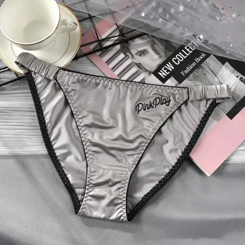 Silver low-rise satin panties with black trim and PinkPlay hip embroidery, flat lay