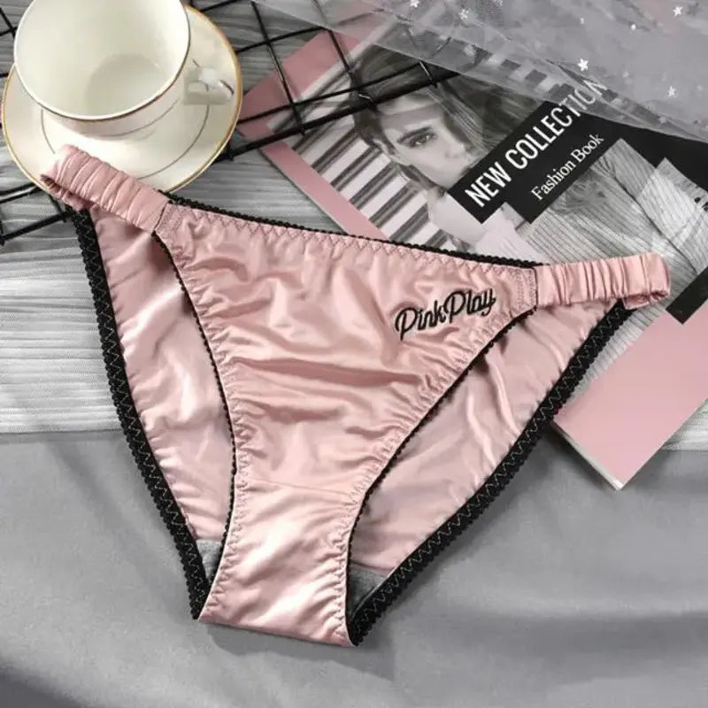 Pink satin bikini panties with black PinkPlay script embroidery and contrast zigzag trim, flat lay on gray fabric beside a fashion book and white teacup