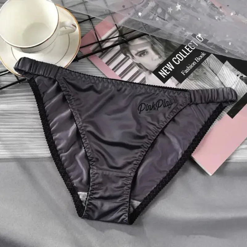 Gray satin bikini panties with black lace trim and embroidered PinkPlay logo on a gray bedsheet flat lay